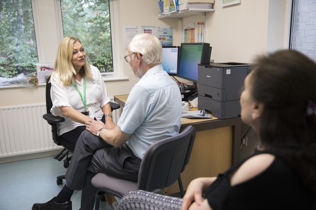 A female practice nurse in a white shirt is with an older patient, a man in a blue shirt.