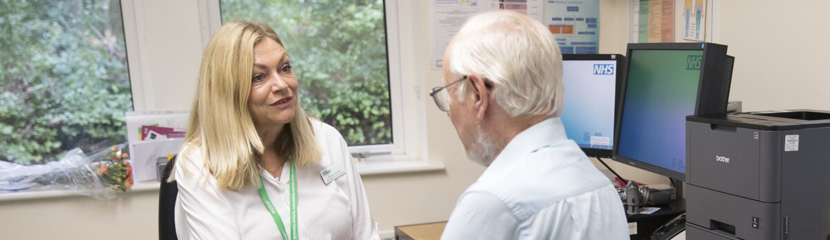A female practice nurse in a white shirt is with an older patient, a man in a blue shirt.