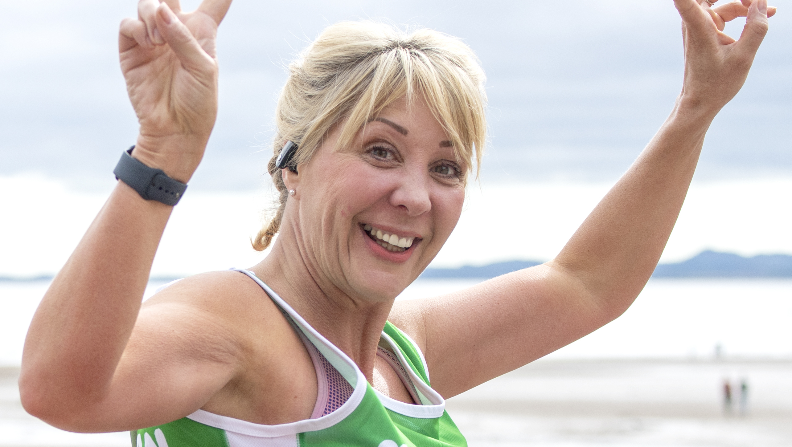Woman wearing a Macmillan t-shirt showing the peace signs and smiling at the camera in front of the beach
