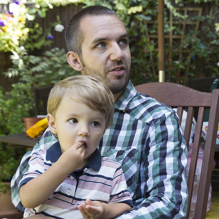 Luke is sat in the garden with his son on his lap.