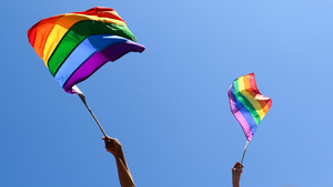 Two rainbow flags being waved against the backdrop of a blue sky.