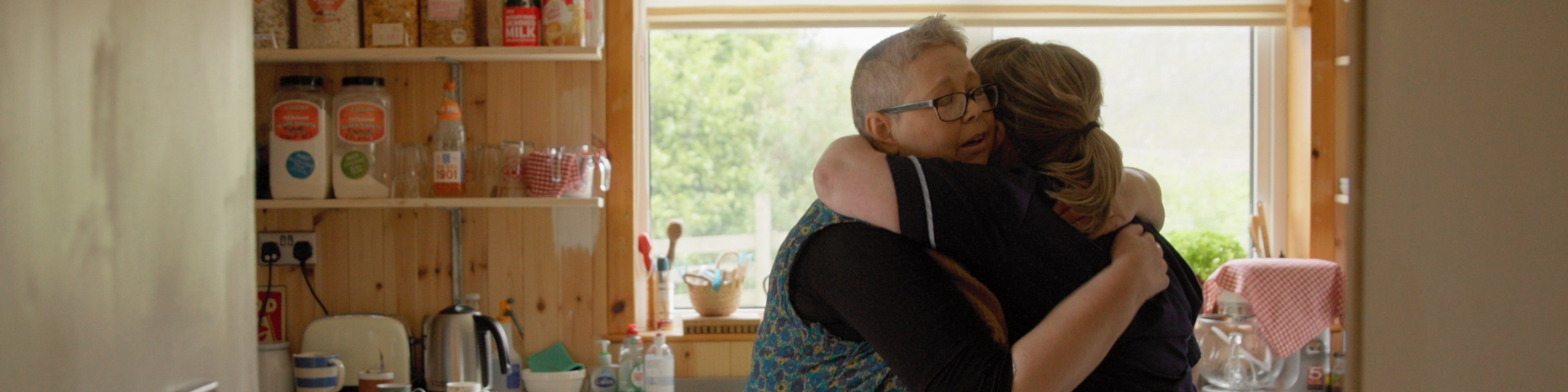 A screenshot from a video created to support the television series, Super Surgeons. Image is of a Macmillan nurse hugging a woman in a kitchen.