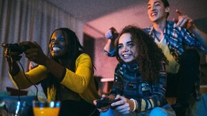 A group of friends sit on a sofa holding controllers and playing computer games