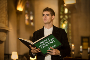 White man in a suit reading a passage from a book at a carol concert 