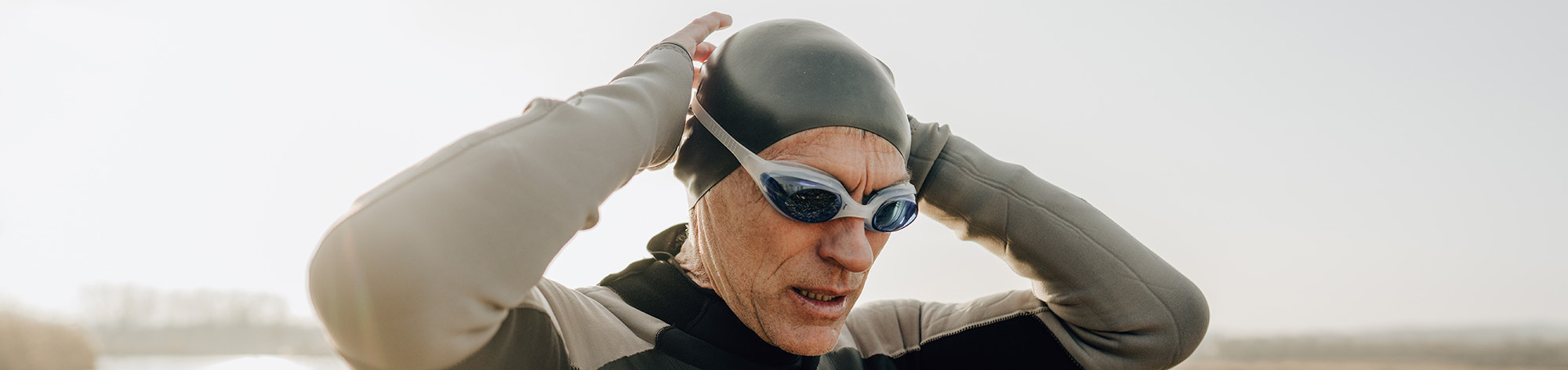 A male swimmer wearing a black swim cap finishes putting on his swimming goggles.