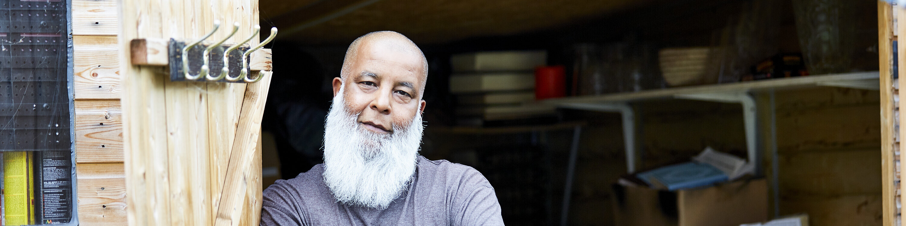 A middle-aged man with a beard and blue t-shirt stands outside a shed.