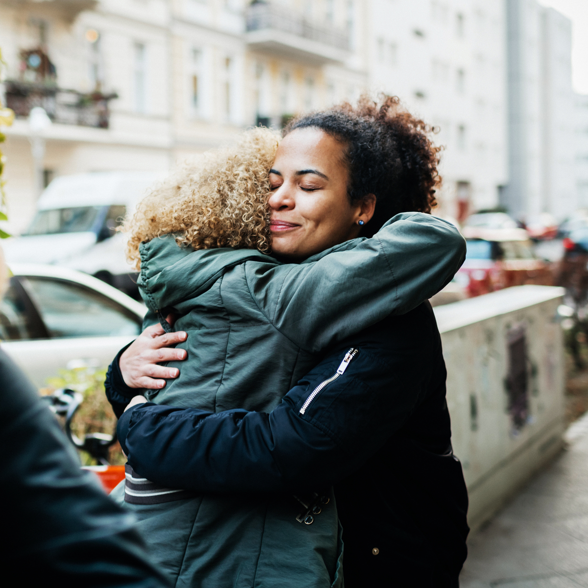 Two women hugging. They are outside on a pavement. They wear coats. One woman faces away from the camera and one woman faces the camera, she is smiling.