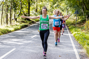 A woman with her arms outstretched running on a road and wearing a green Macmillan running vest