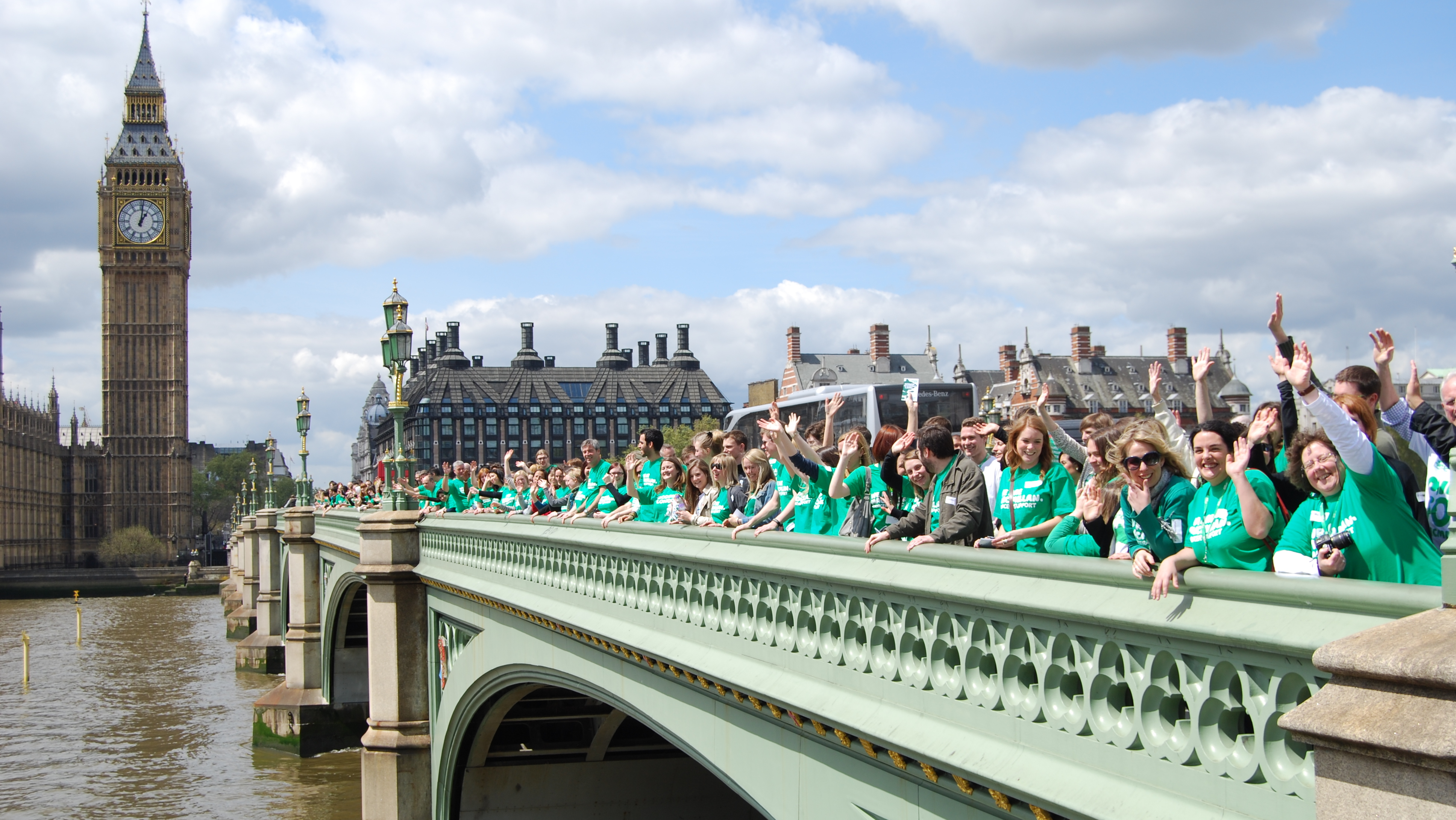 Macmillan staff gathered on Westminster Bridge outside the Houses of Parliament. They are wearing green Macmillan t-shirts. 