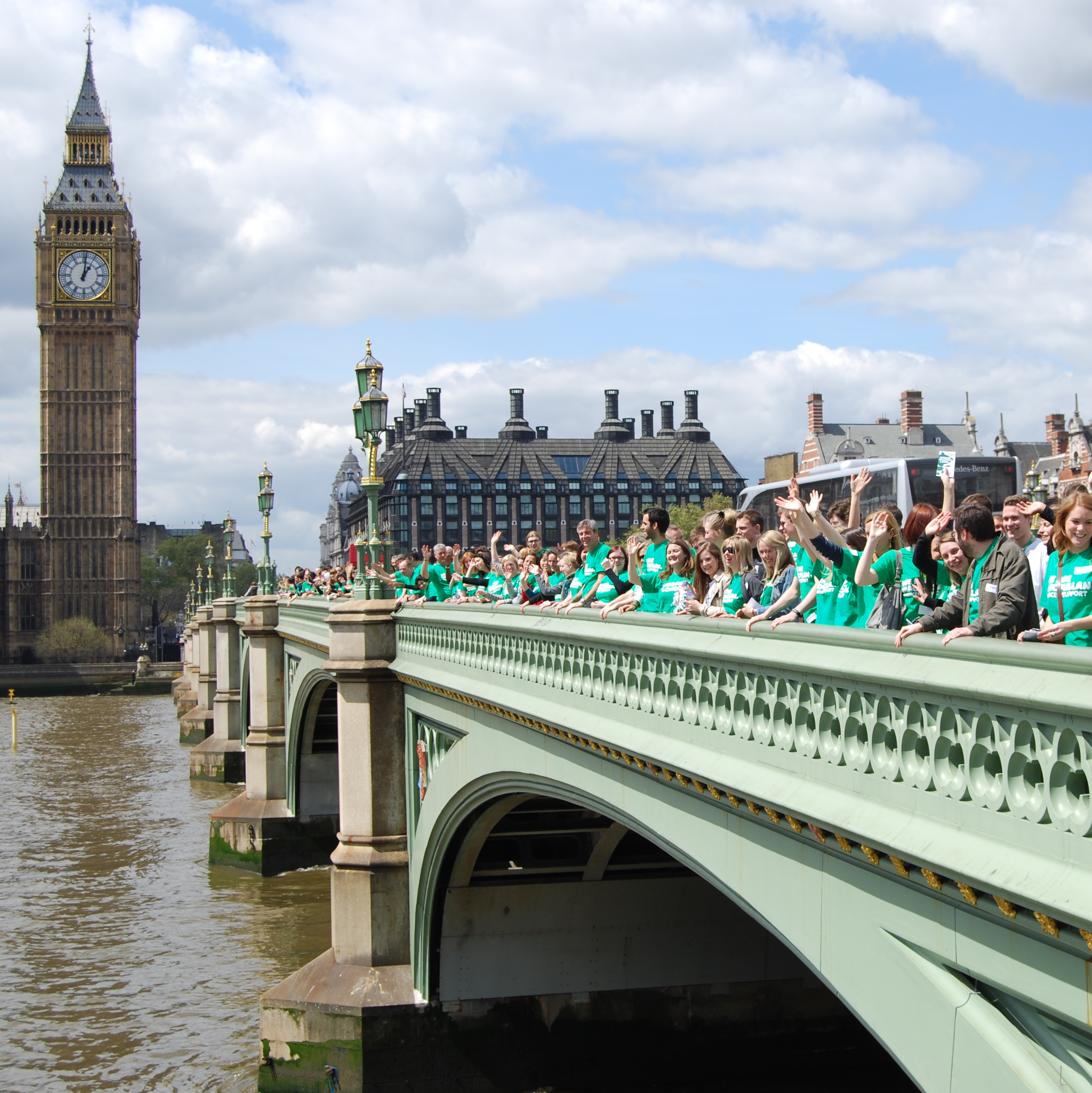 Macmillan staff gathered on Westminster Bridge outside the Houses of Parliament. They are wearing green Macmillan t-shirts. 