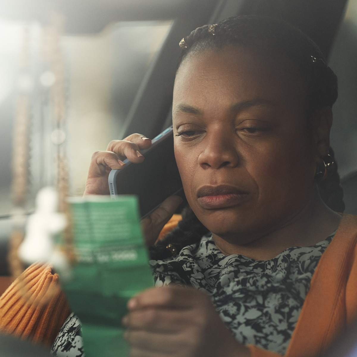Still from a Macmillan TV ad. A person is sitting in their parked car. They are reading a green leaflet and talking on a mobile phone.